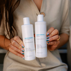 Young dark haired model holding a bottle of ONC MOISTURISING Conditioner 250 mL / 8.4 fl. oz. and a bottle of ONC MOISTURISING Shampoo 250 mL / 8.4 fl. oz. together